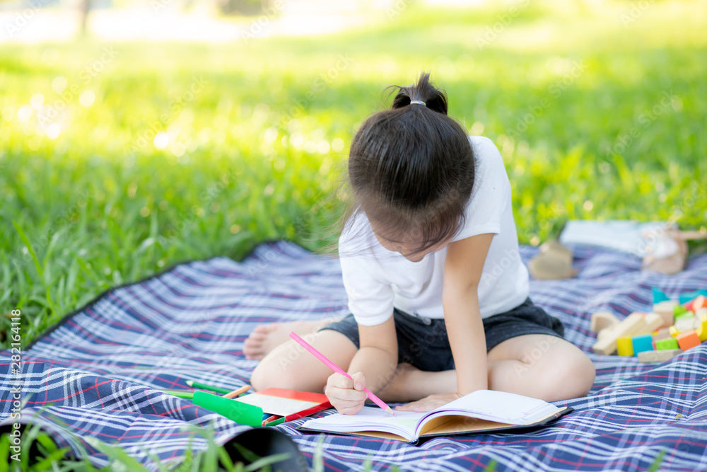 Child sitting on a blanket outdoors, drawing in a book with colorful pencils.