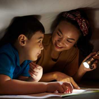 Woman and child lying under a blanket, reading together with a flashlight.