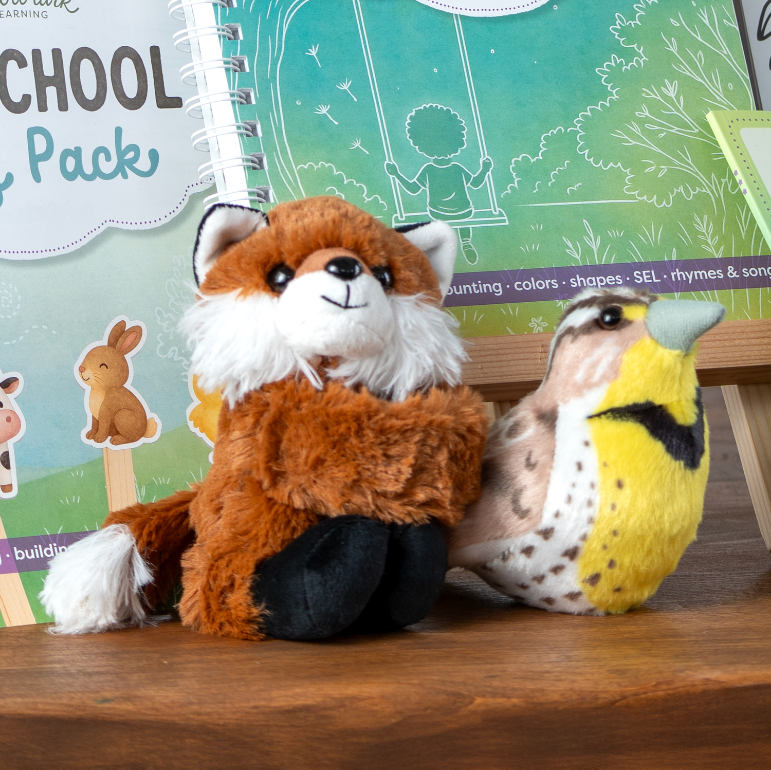 Plush fox and bird toys on a wooden surface with educational materials in the background.