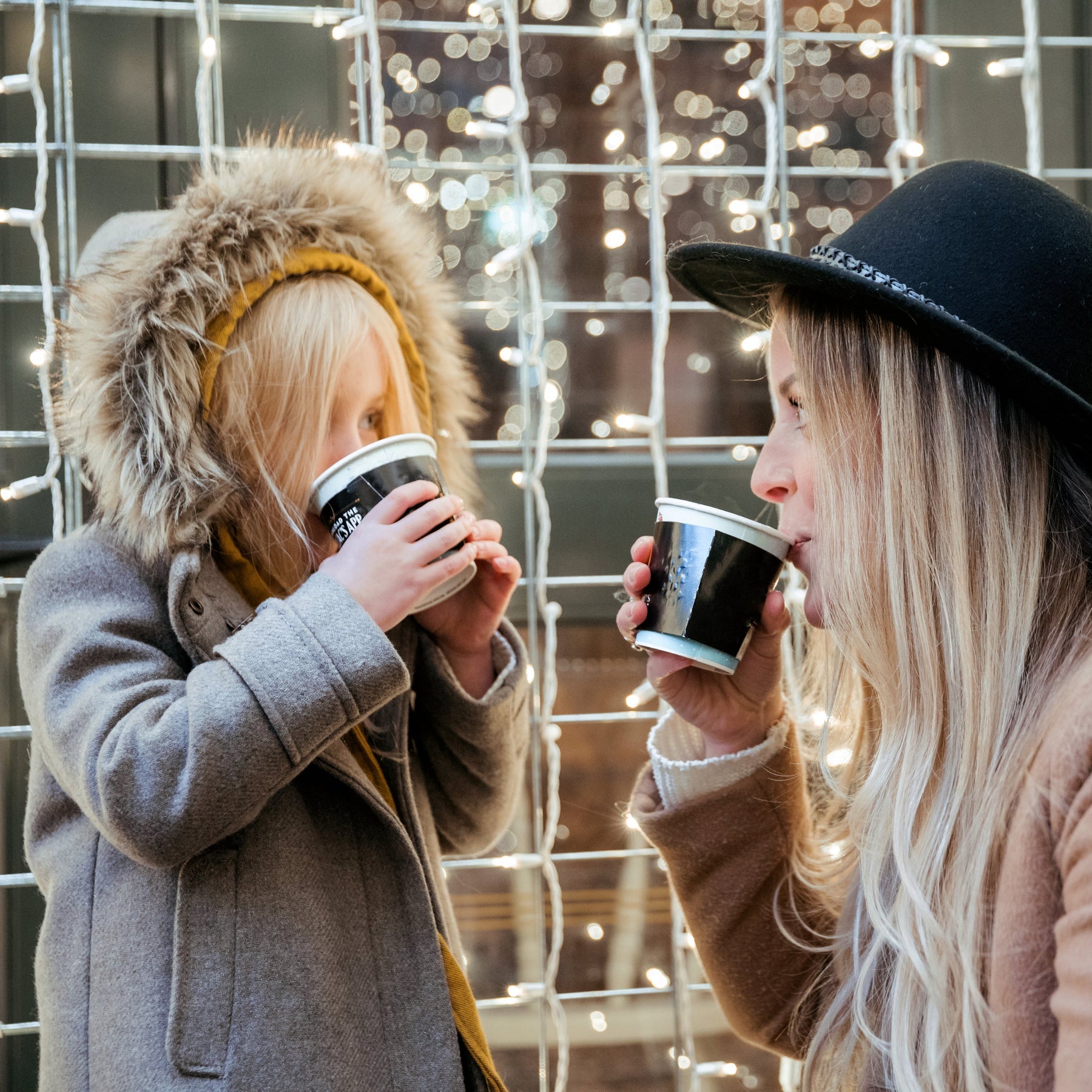 Two people drinking from coffee cups in front of a decorative wire fence with lights.